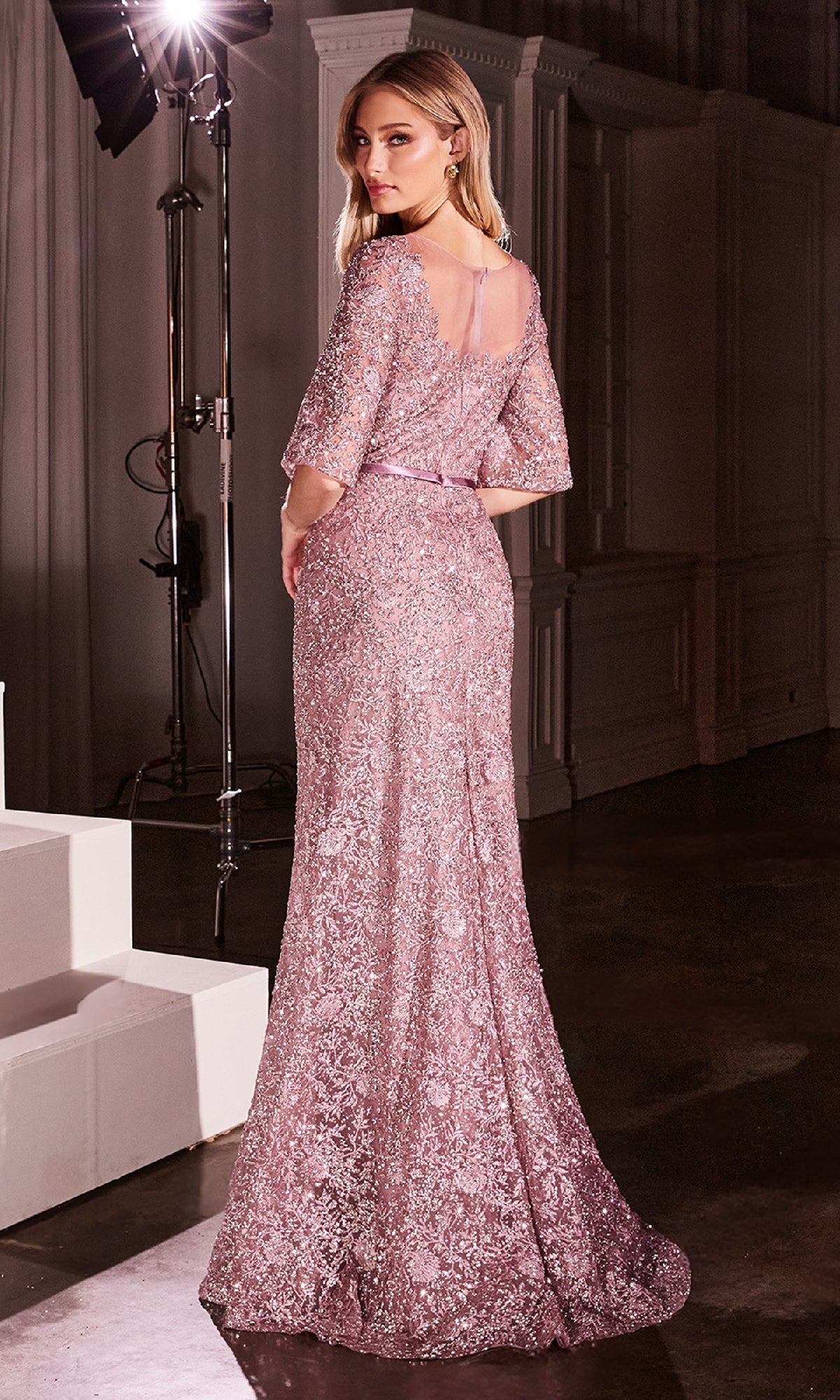 A woman stands on a dark set in the Bell-Sleeve Long Glitter Prom Dress: Ladivine CB149, a floor-length shimmering evening gown with pink floral lace and sheer bell sleeves, looking over her shoulder toward the camera.